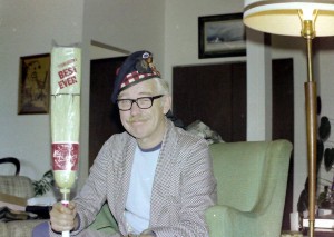 Man in housecoat and pajamas sitting holding a curling broom and wearing a curling hat. Dr. A H Pete Stevens at Christmas with a new curling broom