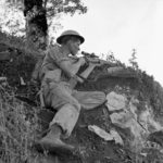 Sniper with helmet and No. 4 MK. I sniper rifle on a hiillside. 1943-10-06 Pte. J. E. (sic) McPhee, a sniper with the Seaforth Highlanders of Canada near Foiano, Italy. The only McPHEE was K74808 Pte. McPHEE, F. J. who served with the Seaforths 21 Aug. 1942 - 26 Dec. 1943. He was wounded 20 Jul. 1943 near Leonforte (West of Mt. Etna), Sicily and was Killed in Action 26 Dec. 1943 in or near Ortona, Italy. He was 23 years old and the husband of Mildred Ethel McPhee of Vancouver, British Columbia, Canada. He is buried in grave VIII. G. 2. Moro River Canadian War Cemetery. (L&AC MIKAN 3207117)