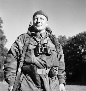 1944-10-06 Cpl. S. Kormendy, a sniper with the Calgary Highlanders at Kapellen, Belgium. Note the captured German 9mm P-38 pistol in the improvised holster and the No. 36 M MK. I fragmentation hand grenade. He is wearing a Denison smock, a face veil as a head scarf, wearing binoculars and has an M1907 leather sling on his No. 4 MK. I (T) sniper rifle. (L&AC MIKAN 3257118)