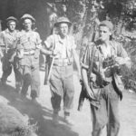 Five soldiers with rifles slubng over their shoulders, walking towards the camera. Lead man is a sniper with binoculars and cigarette. Sniper Canadians head for the Gothic Line in Italy (Hell & High Water)