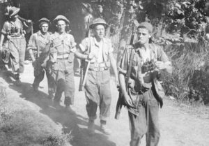 Five soldiers with rifles slubng over their shoulders, walking towards the camera. Lead man is a sniper with binoculars and cigarette. Sniper Canadians head for the Gothic Line in Italy (Hell & High Water)
