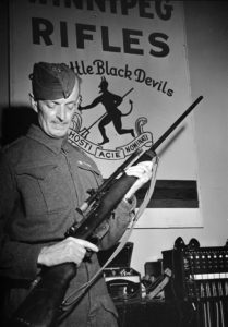 Soldier holding a sniper rifle in an office. 1942-07-30 An unidentified Corporal of the Royal Winnipeg Rifles holding a Ross sniper rifle with a Winchester A5 scope in Ottawa, Ontario. (L&AC PA-208603 MIKAN 3589887)