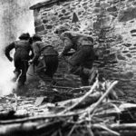 Three Brtiish soldiers rusging a stone building.