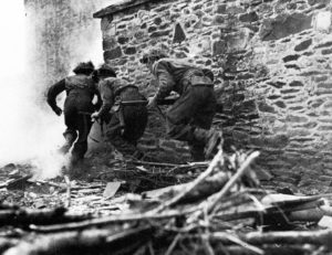 Three Brtiish soldiers rusging a stone building.