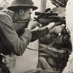 British sniper aiming his sniper rifle through a hole in the wall of a building.