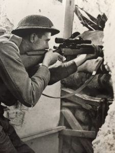 British sniper aiming his sniper rifle through a hole in the wall of a building.