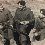 Three men talking with their feet in a trench with a sniper rifle.