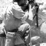 British sniper in Aden in 1964 aiming his No. 4 MK. I (T). He has a Case No. 8 MK. 2 slung to hold the No. 32 MK. 3 scope and its mounting bracket when they are not on the rifle. M1907 leather sling on the rifle. He has an unusual cartridge belt and he also has a clasp knife hanging down on the right side from a lanyard. (TMSS1914 p45)