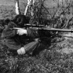 A seated British sniper, legs crossed, resting his elbows on his knees while aiming his sniper rifle.