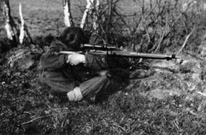 A seated British sniper, legs crossed, resting his elbows on his knees while aiming his sniper rifle.