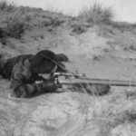 British sniper training among sand dues, lying in a prone position.