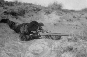 British sniper training among sand dues, lying in a prone position.