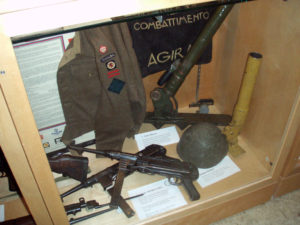 Case 6B - Major Alex "Sandy" Millar's CANLOAN BD blouse and framed CANLOAN scroll; two MP-40 SMGs, Italian helmet and two versions of the 2-INCH mortar. An Italian banner captured at AGIRA is behind them.