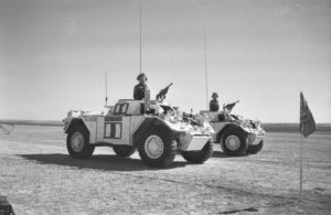 Two white Ferretr Scout Cars driving by with commander standing at attention in each one. United Nations flag in the foreground.
