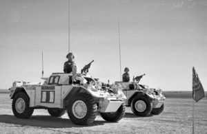 Two white Ferretr Scout Cars driving by with commander standing at attention in each one. United Nations flag in the foreground.